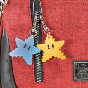 May include: Two star-shaped zipper pulls, one blue and one gold, attached to a red zipper on a red bag. The bag has a black leather trim and a black label with white text that reads "Nintendo Licensed Product Super Mario Bros. Game & Watch".