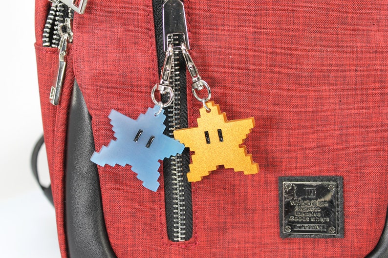 May include: Two star-shaped zipper pulls, one blue and one gold, attached to a red zipper on a red bag. The bag has a black leather trim and a black label with white text that reads "Nintendo Licensed Product Super Mario Bros. Game & Watch".