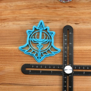 May include: A blue plastic cookie cutter shaped like a stylized crest with a sword and antlers. The cutter is about 2 inches in diameter and is sitting on a wooden surface.