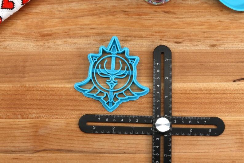 May include: A blue plastic cookie cutter shaped like a stylized crest with a sword and antlers. The cutter is about 2 inches in diameter and is sitting on a wooden surface.