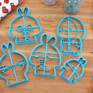 May include: Five blue plastic cookie cutters shaped like cartoon bunnies with different expressions. The cutters are on a wooden surface.