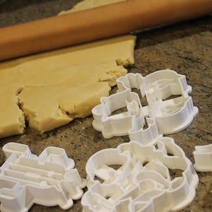 May include: A wooden rolling pin rolls out dough on a speckled countertop. Several white plastic cookie cutters in various shapes are scattered nearby, ready for use in baking. The dough is a light tan color.