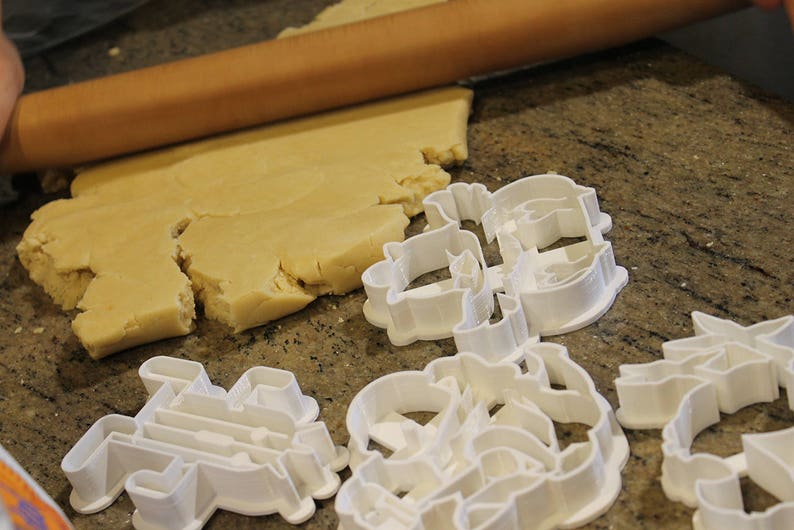 May include: A wooden rolling pin rolls out dough on a speckled countertop. Several white plastic cookie cutters in various shapes are scattered nearby, ready for use in baking. The dough is a light tan color.
