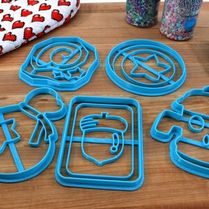 May include: A set of six blue plastic cookie cutters shaped like a star, a bag, an acorn, a swirl, a robot, and a shield.