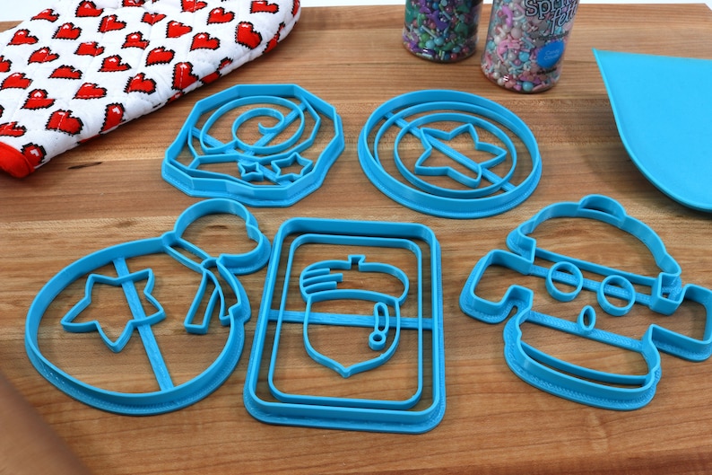 May include: A set of six blue plastic cookie cutters shaped like a star, a bag, an acorn, a swirl, a robot, and a shield.