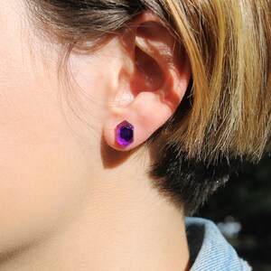 May include: A close-up of a person's ear with a purple gemstone stud earring. The earring is a hexagonal shape with a faceted design.