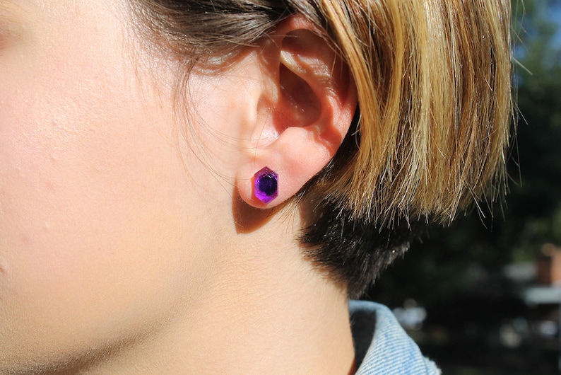May include: A close-up of a person's ear with a purple gemstone stud earring. The earring is a hexagonal shape with a faceted design.