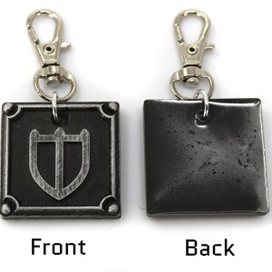 May include: A silver metal keychain with a square charm featuring a shield design on the front and a plain surface on the back. The keychain has a silver metal clasp.