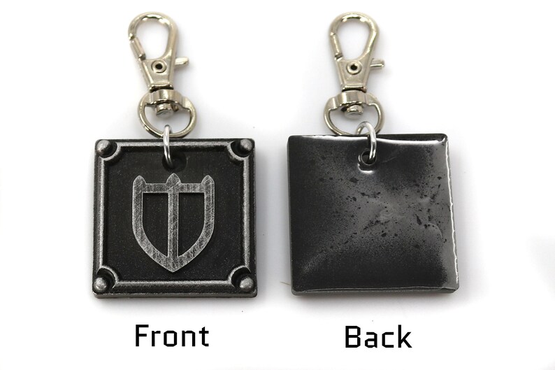 May include: A silver metal keychain with a square charm featuring a shield design on the front and a plain surface on the back. The keychain has a silver metal clasp.