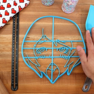 May include: A blue plastic cookie cutter shaped like a cartoon face with a grid pattern. The cutter is on a wooden surface with a rolling pin, a ruler, a spatula, and a red oven mitt.