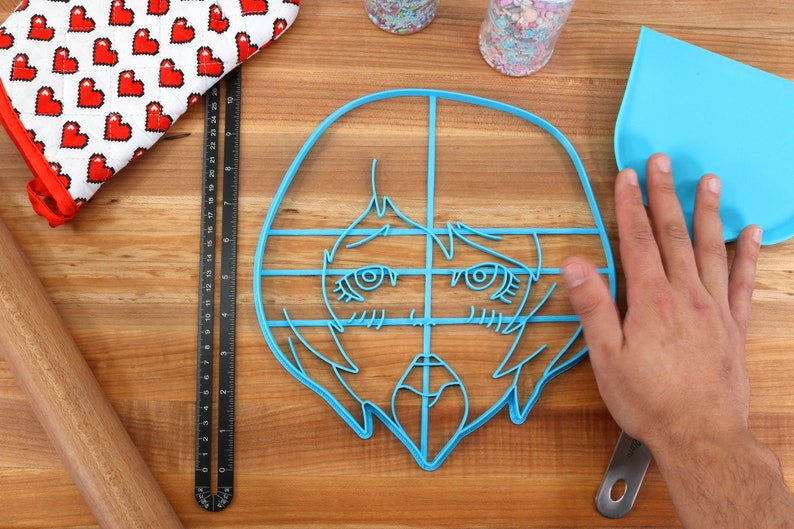 May include: A blue plastic cookie cutter shaped like a cartoon face with a grid pattern. The cutter is on a wooden surface with a rolling pin, a ruler, a spatula, and a red oven mitt.