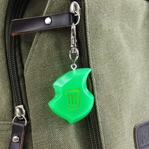 May include: A green plastic keychain charm shaped like a leaf with a harp symbol inside. The charm is attached to a silver chain and is hanging from a green canvas bag.