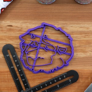 May include: Purple plastic cookie cutter shaped like a cartoon face with a wide smile and a mustache. The cookie cutter is on a wooden surface.