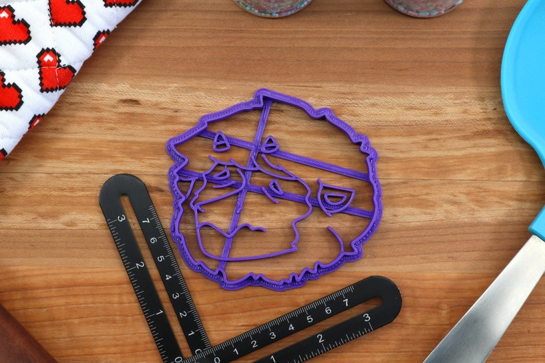 May include: Purple plastic cookie cutter shaped like a cartoon face with a wide smile and a mustache. The cookie cutter is on a wooden surface.
