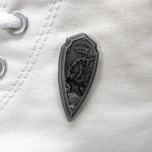 May include: A silver-colored shield-shaped pin with a black and white image of a mythical creature with a crown on top.