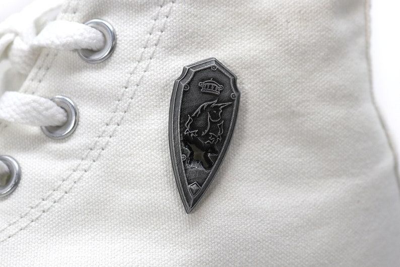 May include: A silver-colored shield-shaped pin with a black and white image of a mythical creature with a crown on top.