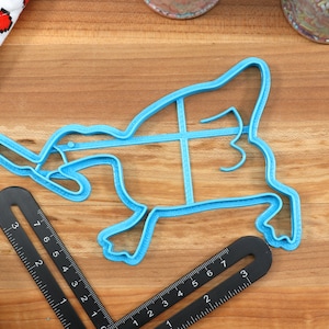 May include: A blue plastic cookie cutter shaped like a duck. The cutter is on a wooden surface with a ruler next to it.