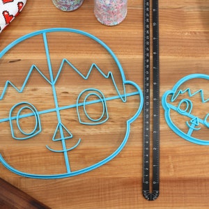 May include: Two blue plastic cookie cutters shaped like a person's face. The larger cutter has a grid pattern inside. The smaller cutter is a smaller version of the larger cutter.