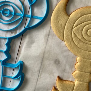 Op de afbeelding: Een blauwe plastic koekjesvormer en een lichtbruine suikerkoek, beide in de vorm van een sleutel met een oogontwerp in het midden.