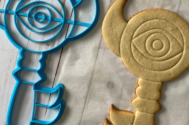 Op de afbeelding: Een blauwe plastic koekjesvormer en een lichtbruine suikerkoek, beide in de vorm van een sleutel met een oogontwerp in het midden.