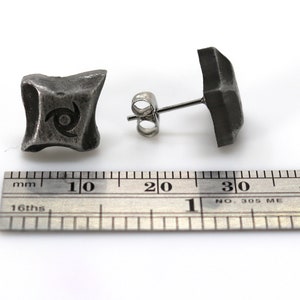 Puede incluir: Dos pendientes de bot&oacute;n cuadrados de color gris oscuro. Un pendiente muestra un dise&ntilde;o en espiral. Los pendientes est&aacute;n sobre una superficie blanca junto a una regla. Los pendientes son de metal.