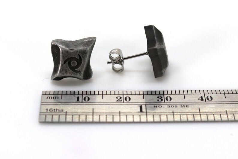 Puede incluir: Dos pendientes de bot&oacute;n cuadrados de color gris oscuro. Un pendiente muestra un dise&ntilde;o en espiral. Los pendientes est&aacute;n sobre una superficie blanca junto a una regla. Los pendientes son de metal.