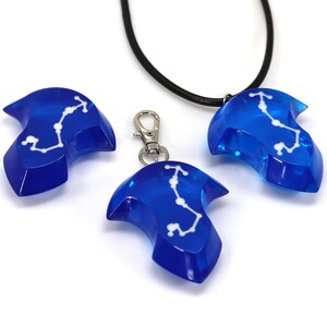 May include: Three blue resin pendants with a white constellation design. One pendant is on a black cord necklace, one is on a silver chain, and one is on a silver key chain.