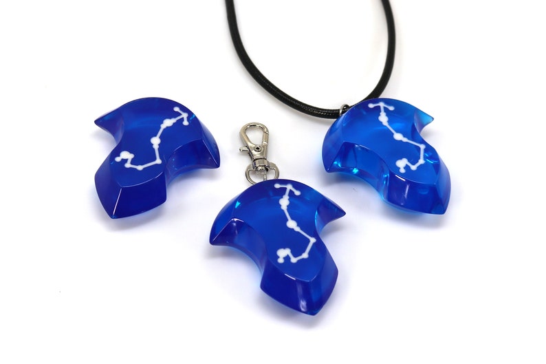 May include: Three blue resin pendants with a white constellation design. One pendant is on a black cord necklace, one is on a silver chain, and one is on a silver key chain.
