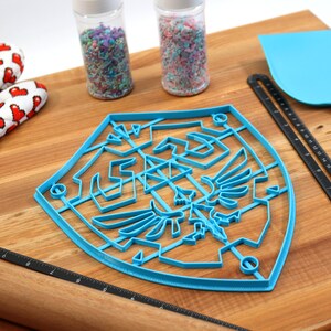 May include: Blue plastic cookie cutter shaped like the Triforce symbol from the Legend of Zelda video game series. The cutter is on a wooden surface with a rolling pin, a spatula, and two jars of sprinkles.