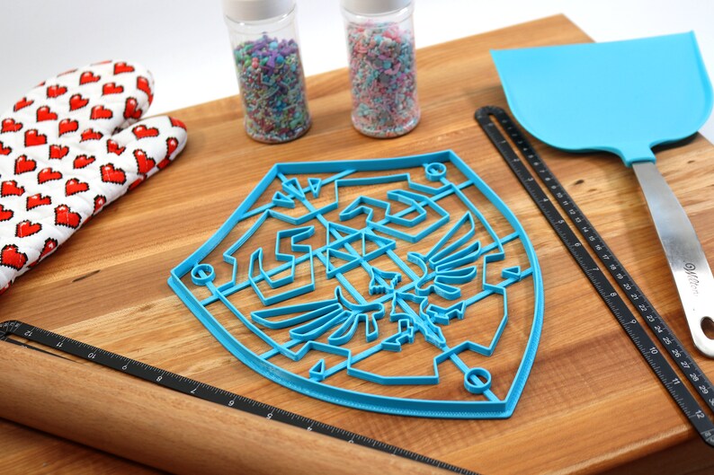 May include: Blue plastic cookie cutter shaped like the Triforce symbol from the Legend of Zelda video game series. The cutter is on a wooden surface with a rolling pin, a spatula, and two jars of sprinkles.