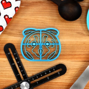 May include: Blue plastic cookie cutter shaped like a raccoon face. The cutter has a detailed design with eyes, nose, and ears.