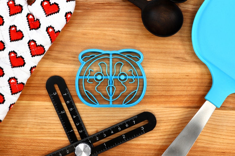 May include: Blue plastic cookie cutter shaped like a raccoon face. The cutter has a detailed design with eyes, nose, and ears.