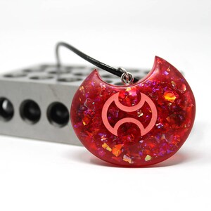May include: A red crescent moon pendant necklace with a white symbol inside. The pendant is made of resin and has a glittery finish.