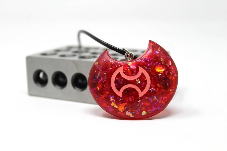 May include: A red crescent moon pendant necklace with a white symbol inside. The pendant is made of resin and has a glittery finish.