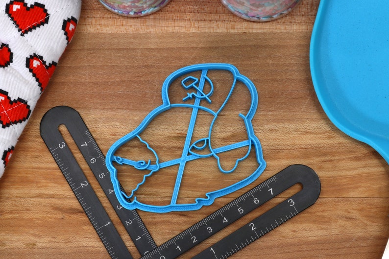 May include: A blue plastic cookie cutter shaped like a cartoon character wearing a hat with a hammer and sickle symbol.