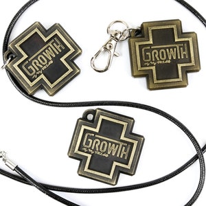 May include: Two gold-colored metal cross charms with the word "GROWTH" on them. One charm is on a black cord necklace and the other is on a silver key chain.