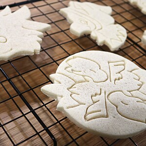May include: Five plain sugar cookies on a black wire cooling rack. The cookies are in the shapes of a dragon, a snowflake, and three different types of birds.