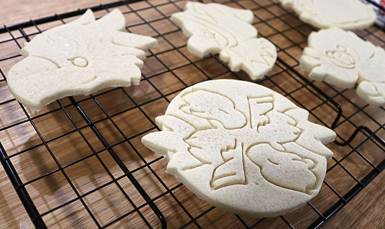 May include: Five plain sugar cookies on a black wire cooling rack. The cookies are in the shapes of a dragon, a snowflake, and three different types of birds.
