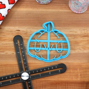 May include: A blue plastic pumpkin cookie cutter with a "uwu" face. The cutter is on a wooden surface.