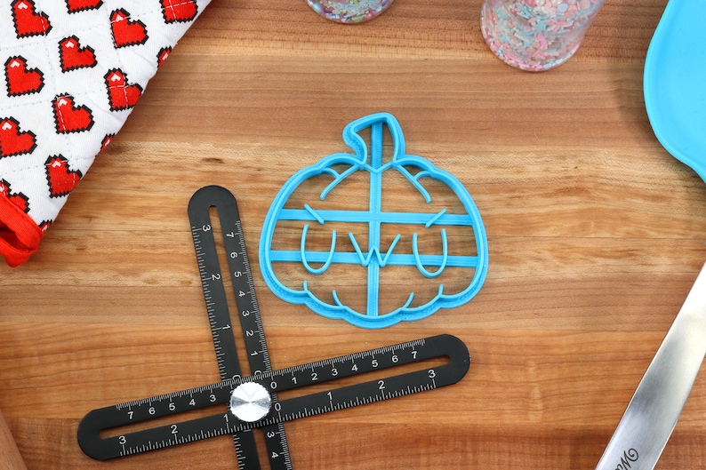 May include: A blue plastic pumpkin cookie cutter with a "uwu" face. The cutter is on a wooden surface.