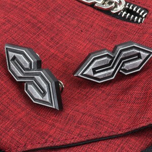May include: Two geometric, silver-colored metal pins with a black outline, shaped like stylized letter "S" designs. The pins are set against a textured red fabric background, with a silver zipper visible.