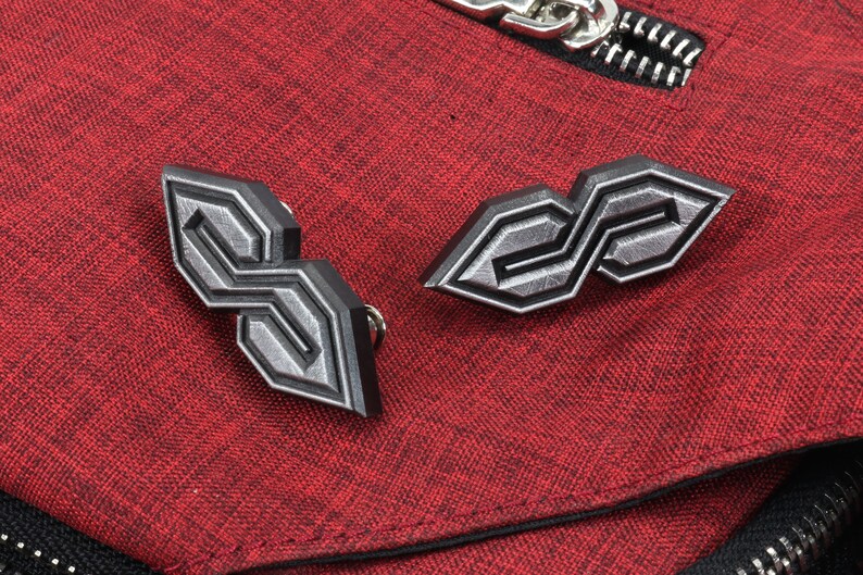 May include: Two geometric, silver-colored metal pins with a black outline, shaped like stylized letter "S" designs. The pins are set against a textured red fabric background, with a silver zipper visible.