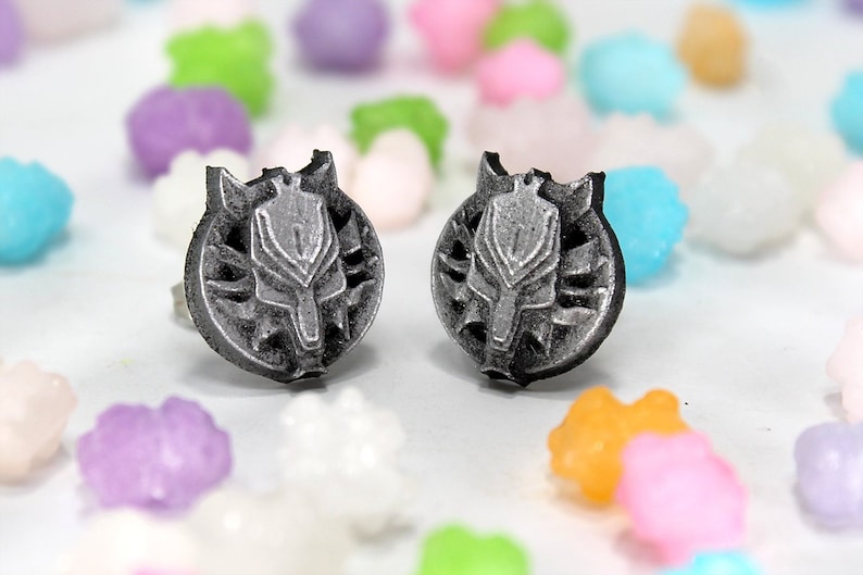 Op de afbeelding: Een paar zilverkleurige oorbel studs met een wolfskop ontwerp. De oorbellen zijn geplaatst tegen een witte achtergrond met kleurrijk snoep.