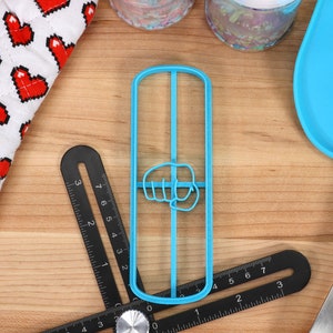 May include: A blue plastic cookie cutter shaped like a fist with the thumb pointing to the left. The cookie cutter is on a wooden surface with a ruler and a blue silicone spatula.