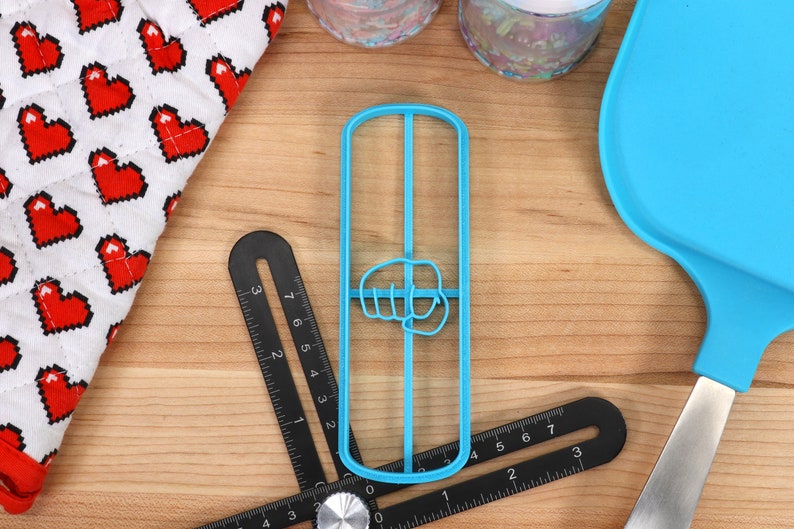 May include: A blue plastic cookie cutter shaped like a fist with the thumb pointing to the left. The cookie cutter is on a wooden surface with a ruler and a blue silicone spatula.