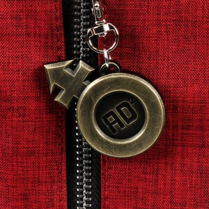 May include: A red fabric zipper pull with a gold metal charm. The charm has a circular design with the letters "AD" in the center. The charm is attached to a small gold metal arrow.