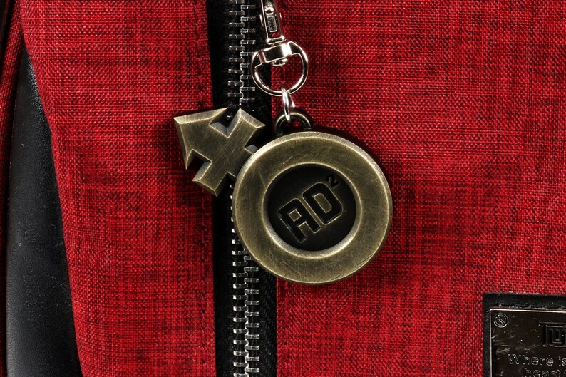May include: A red fabric zipper pull with a gold metal charm. The charm has a circular design with the letters "AD" in the center. The charm is attached to a small gold metal arrow.