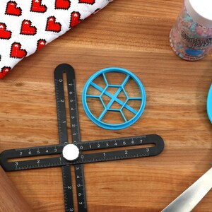 May include: A blue plastic cookie cutter shaped like a honeycomb, a black ruler with a white knob, a wooden rolling pin, a blue spatula with a silver handle, and a jar of colorful sprinkles.