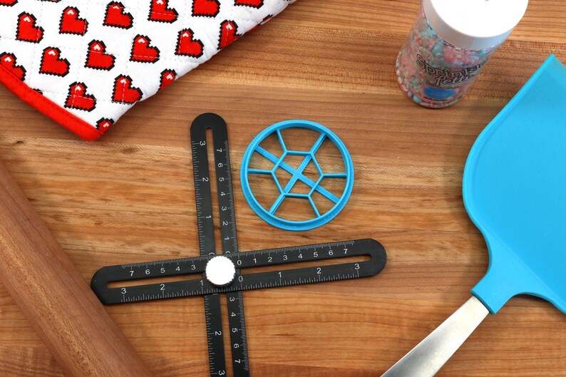 May include: A blue plastic cookie cutter shaped like a honeycomb, a black ruler with a white knob, a wooden rolling pin, a blue spatula with a silver handle, and a jar of colorful sprinkles.