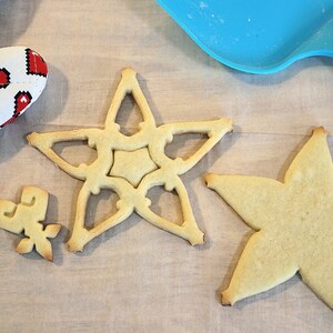 Pu&ograve; includere: Vista dall'alto di biscotti a forma di stella di varie dimensioni, accanto a un guanto da forno rosso e bianco con un motivo a cuore e una spatola blu. I biscotti sono dorati e il guanto ha uno sfondo bianco con cuori pixelati rossi.
